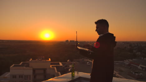 A-male-stockbroker-freelancer-stands-on-a-rooftop-at-sunset-with-a-laptop-and-types-on-a-keyboard-with-his-fingers-looking-at-the-cityscape-from-a-bird's-eye-view.